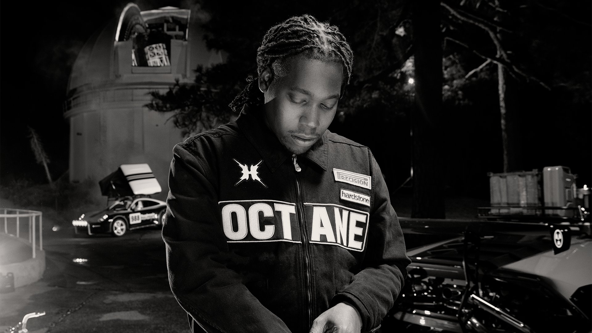 Schwarz‑weißes Foto von Don Toliver in „OCTANE“-Jacke, der nachts neben Autos und einem runden Gebäude nach unten schaut.