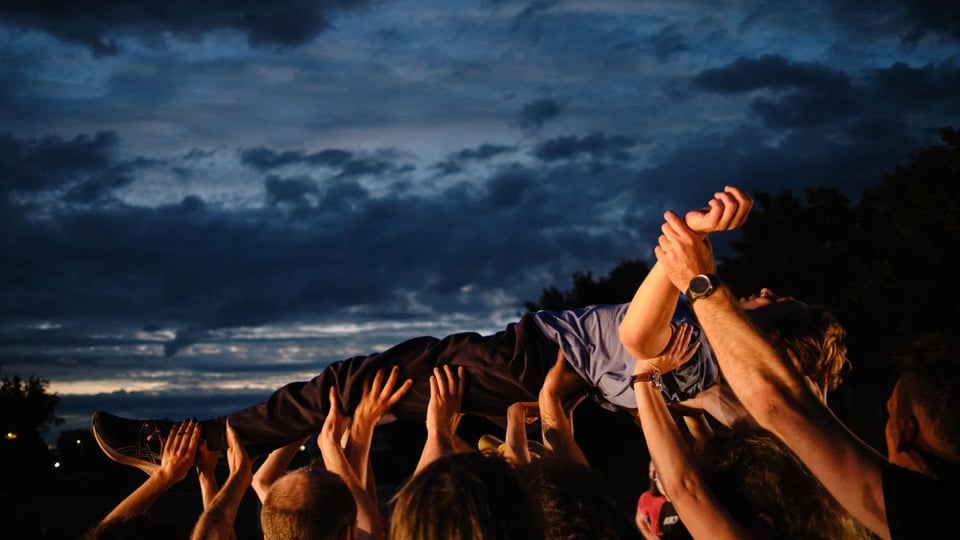 Eine Person wird im Freien von vielen erhobenen Händen über die Menge getragen, vor dunklem Abendhimmel.