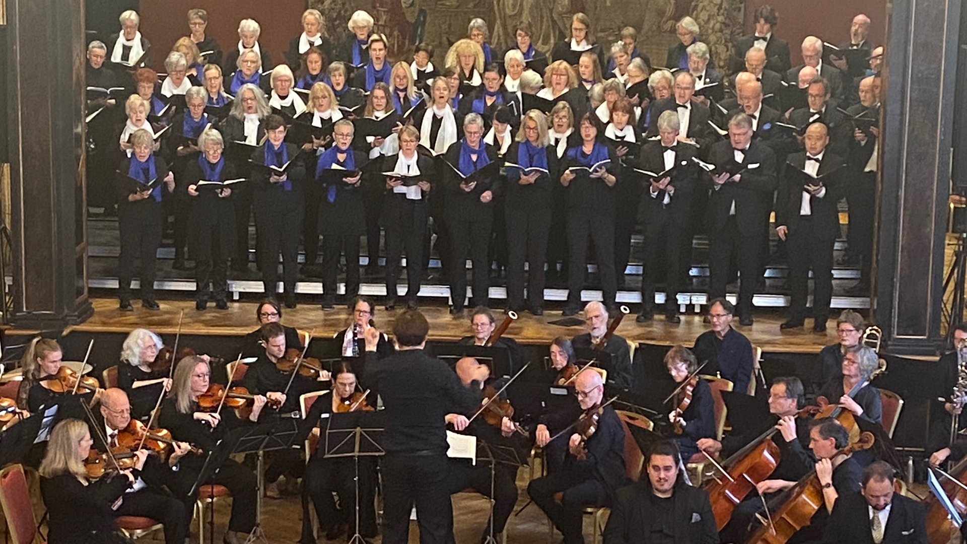 Konzertaufführung mit großem Chor auf gestuften Podesten und Sinfonieorchester im Vordergrund. Die Mitwirkenden tragen schwarze Konzertkleidung mit weißen und blauen Schals bzw. Fliegen; in der Mitte dirigiert eine Person das Ensemble