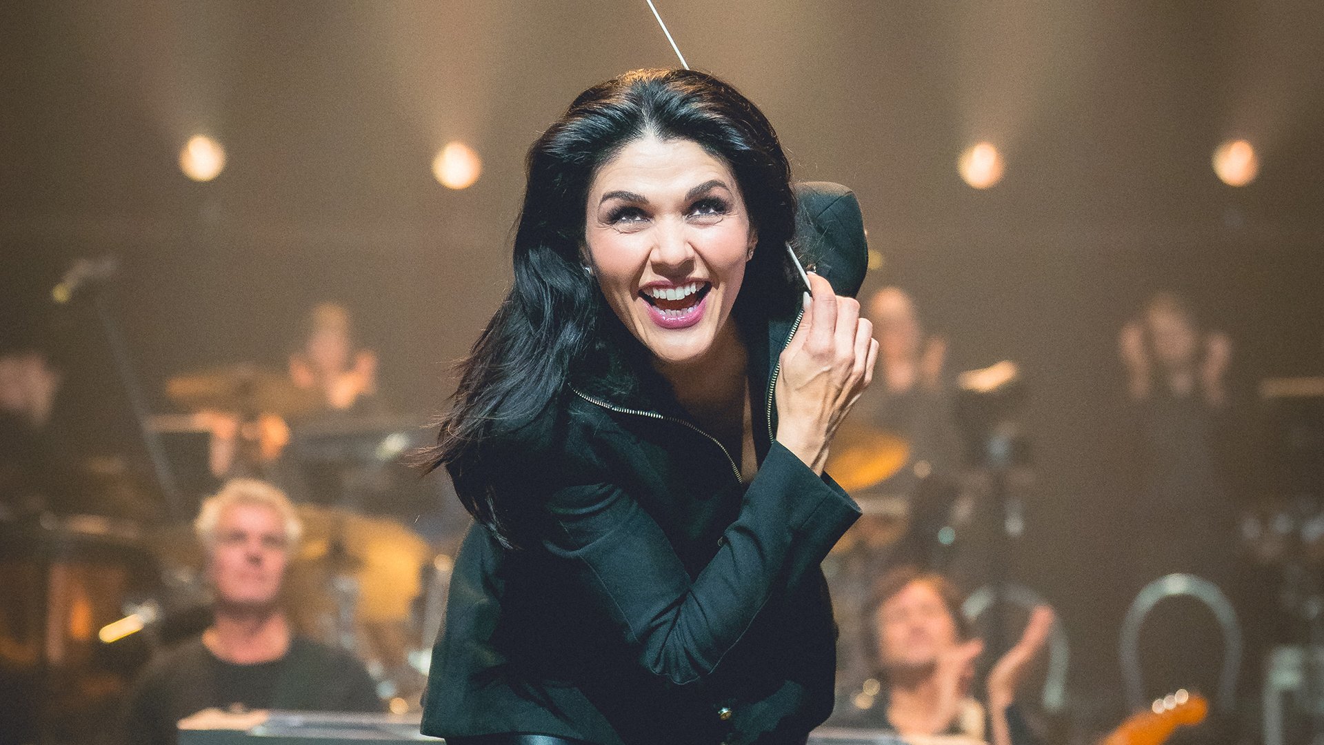 Alexandra Arrieche in schwarzem Blazer mit Dirigentenstock, im Hintergrund weitere Musiker