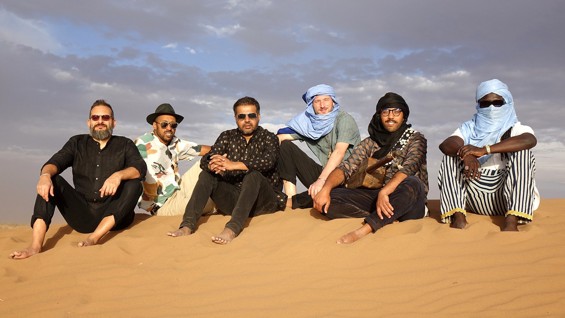 Foto für die Veranstaltung „JISR & GUESTS: Klänge der Sehnsucht – Von der Sahara bis München“, mit den Künstlern, die auf einer Düne in der Sahara sitzen.