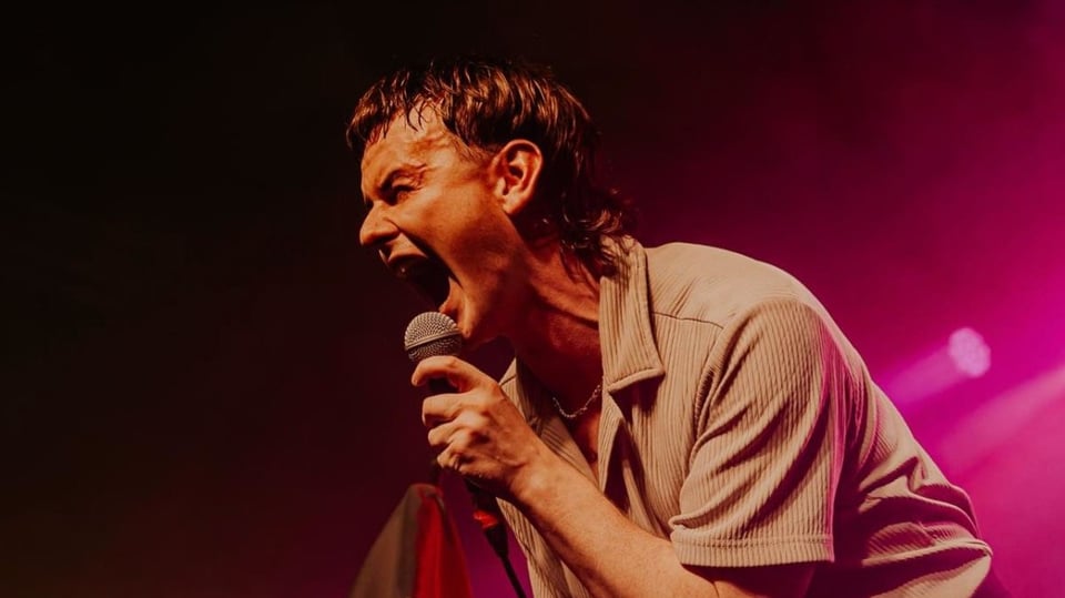 Der Künstler Declan Welsh in heller Kleidung live mit einem Mikrofon in pinkem und dunkelrotem Scheinwerferlicht 