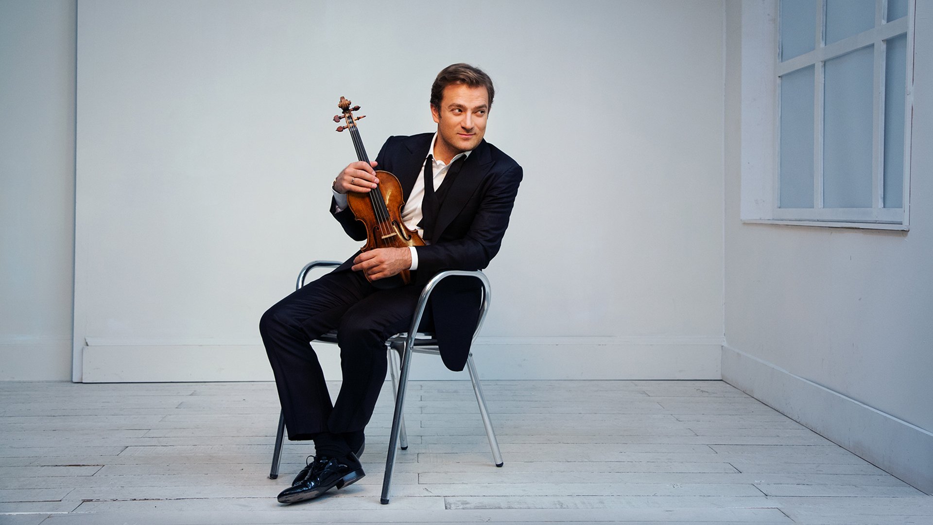 Renaud Capuçon sitzt in einem hellen, minimalistischen Raum auf einem Stuhl und hält eine Violine, trägt einen dunklen Anzug, neben ihm ein Fenster und heller Holzboden