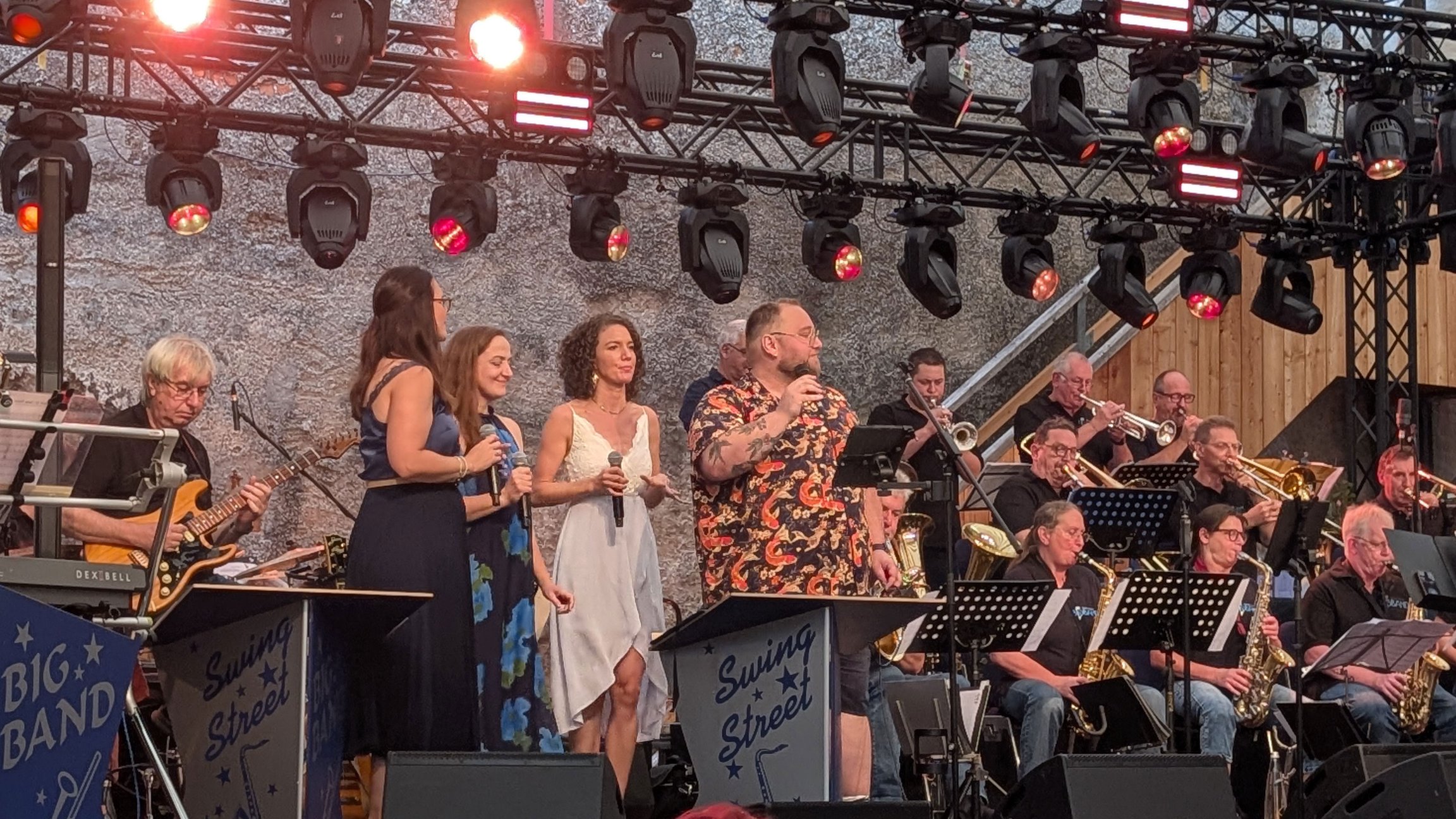 Big Band mit Sängerinnen, Sänger und Bläsern bei einem Konzert auf einer beleuchteten Bühne. Auf den Notenpulten stehen die Aufschriften Big Band und Swing Street.