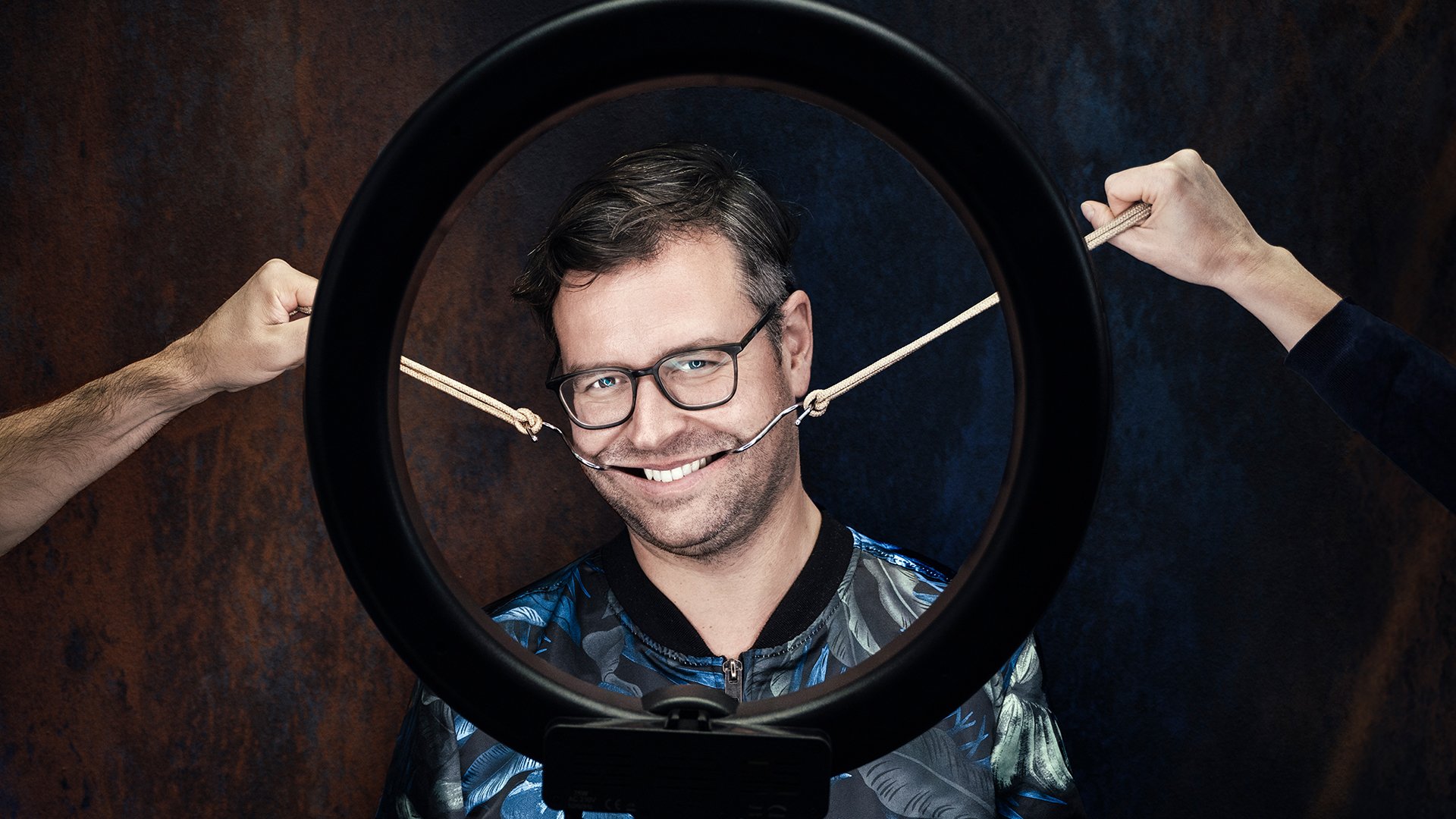 Der Künstler hinter einem kreisförmigen Studiolicht mit zwei Armen links und rechts, die mit Haken seinen Mund auseinanderziehen