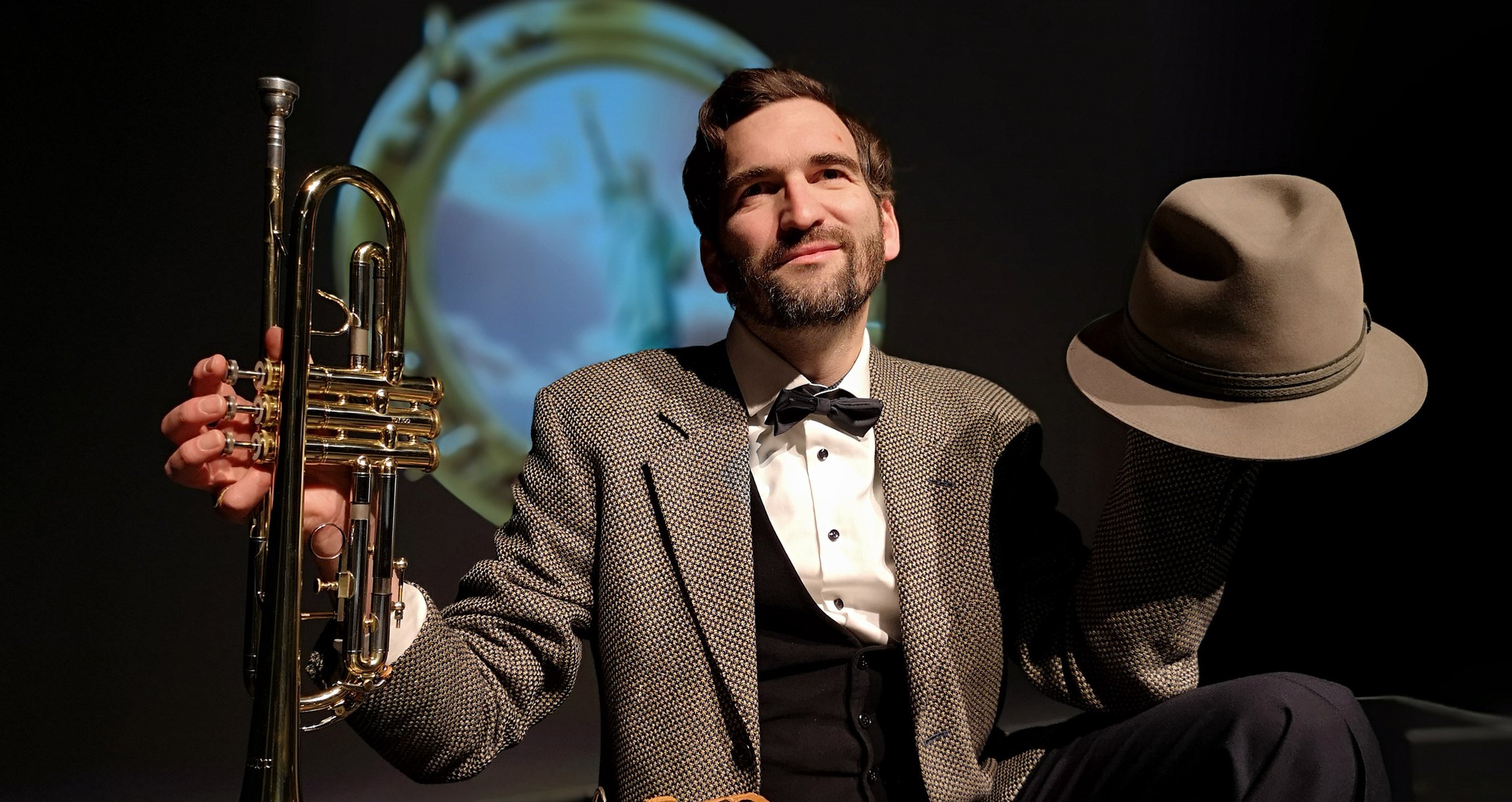 Mann in braunem Sakko und Fliege mit Trompete und Hut in der Hand