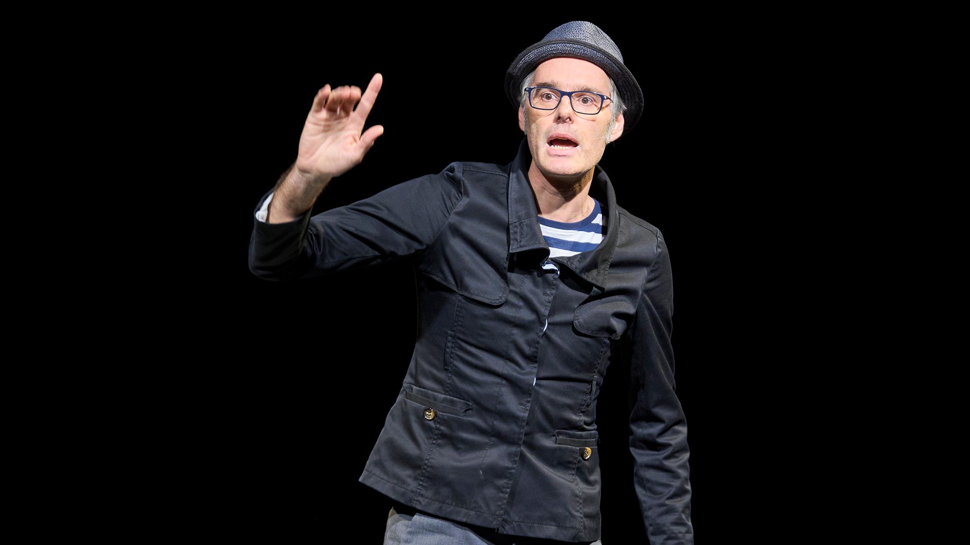 Ein Darsteller mit schwarzer Jacke und hut auf einer Bühne