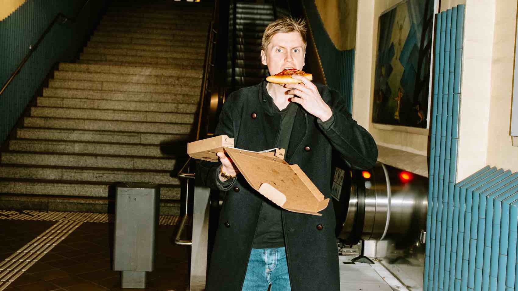 Till Reiners steht vor einer Rolltreppe und isst ein Stück Pizza