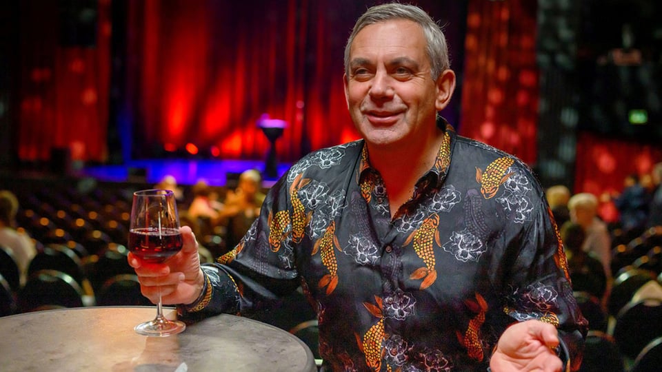 Porträtfoto von Wladimir Kaminer an einem Stehtisch mit einem Glas Rotwein in der Hand, vor einem Veranstaltungsraum mit Reihen von Stühlen und rot beleuchteter Bühne im Hintergrund