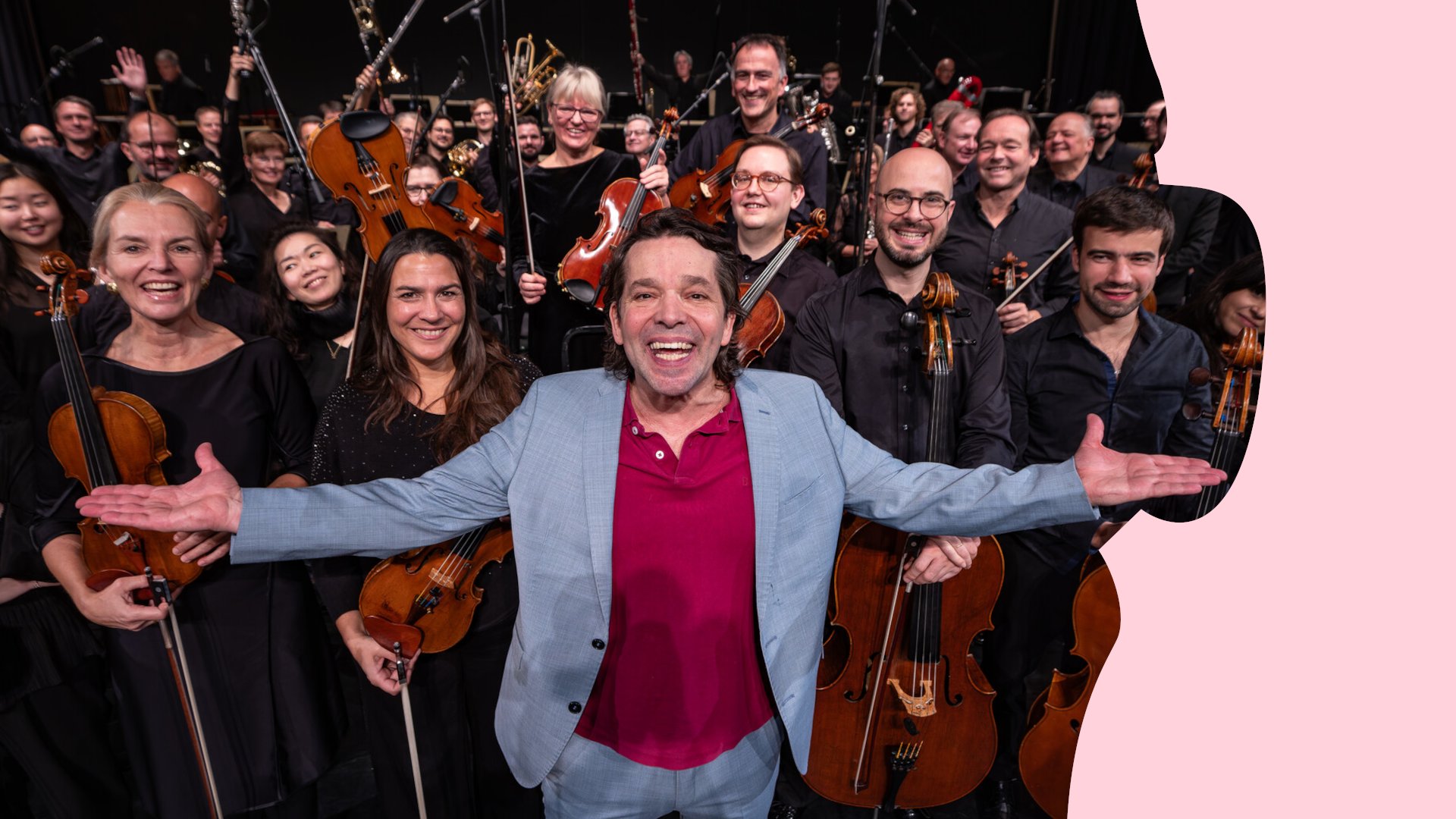 Person mit ausgebreiteten Armen vor einem Orchester; die Musikerinnen und Musiker halten Streich- und Blasinstrumente und blicken in die Kamera.