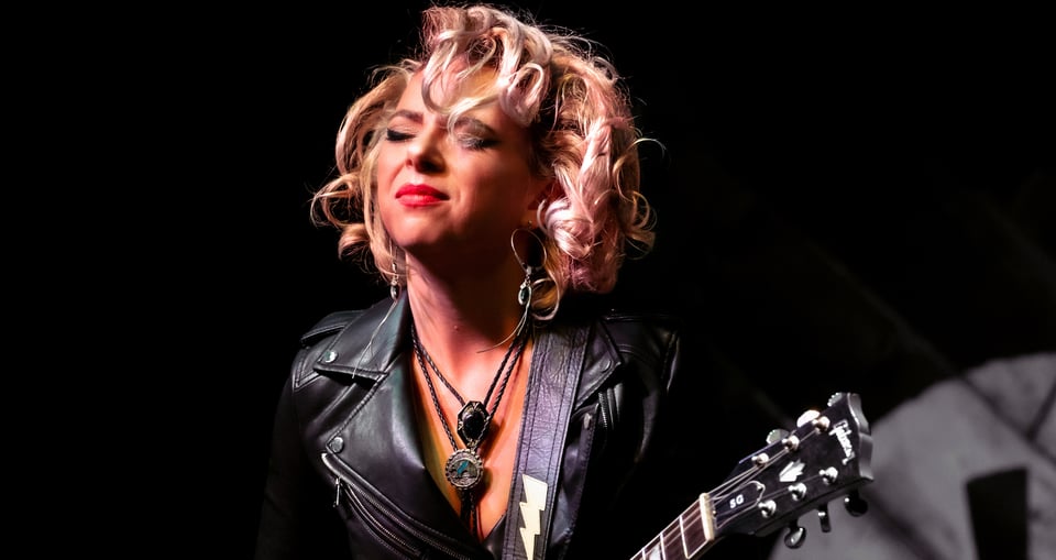Blonde Frau in schwarzer Lederjacke mit Gitarre in der Hand