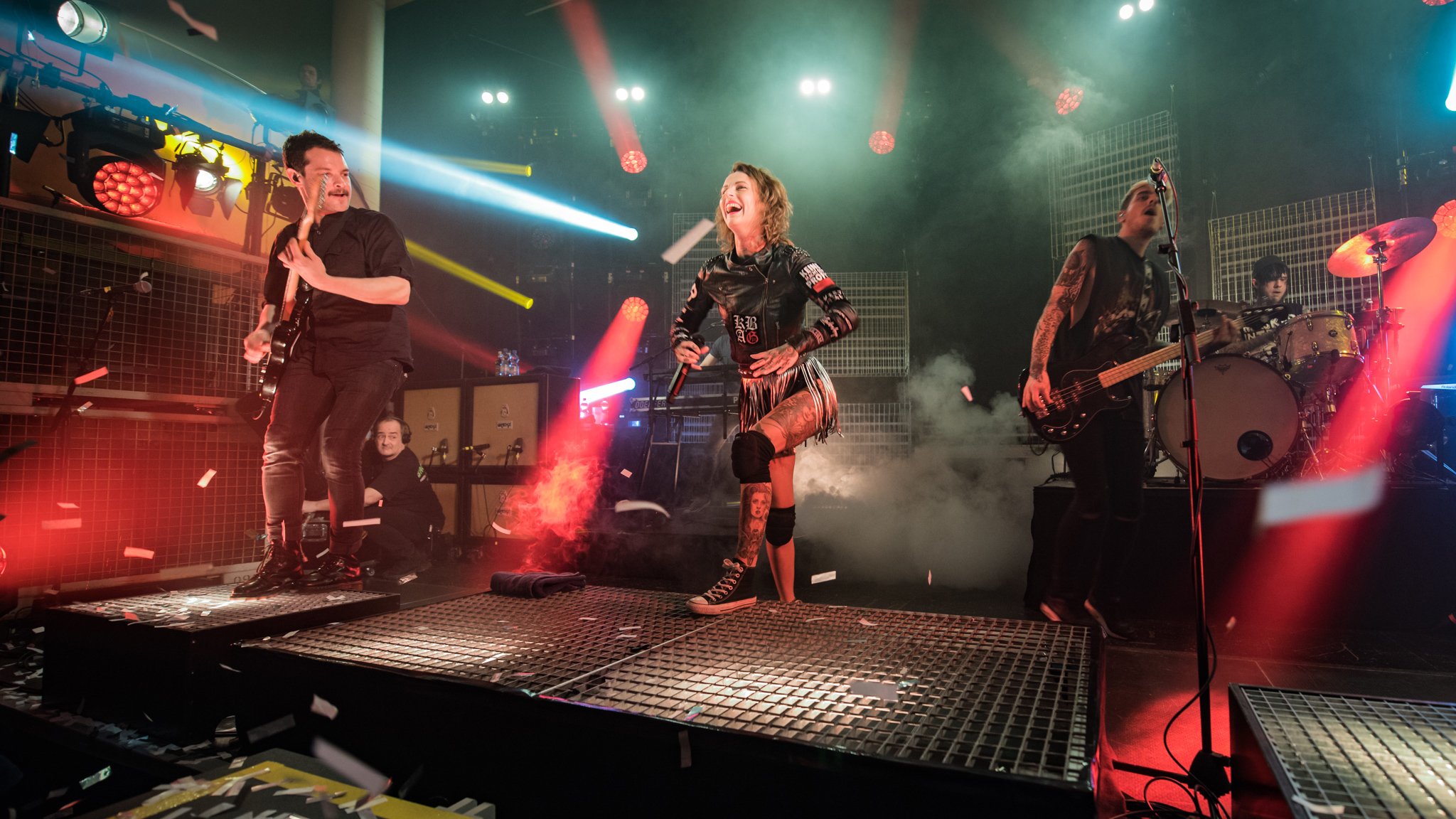 Bühnenfoto von Jennifer Rostock mit Sängerin und Band bei einem Liveauftritt auf einer Konzertbühne, umgeben von Nebel, Konfetti und rot blauem Scheinwerferlicht, mit Gitarrist, Bassist und Schlagzeuger im Hintergrund.