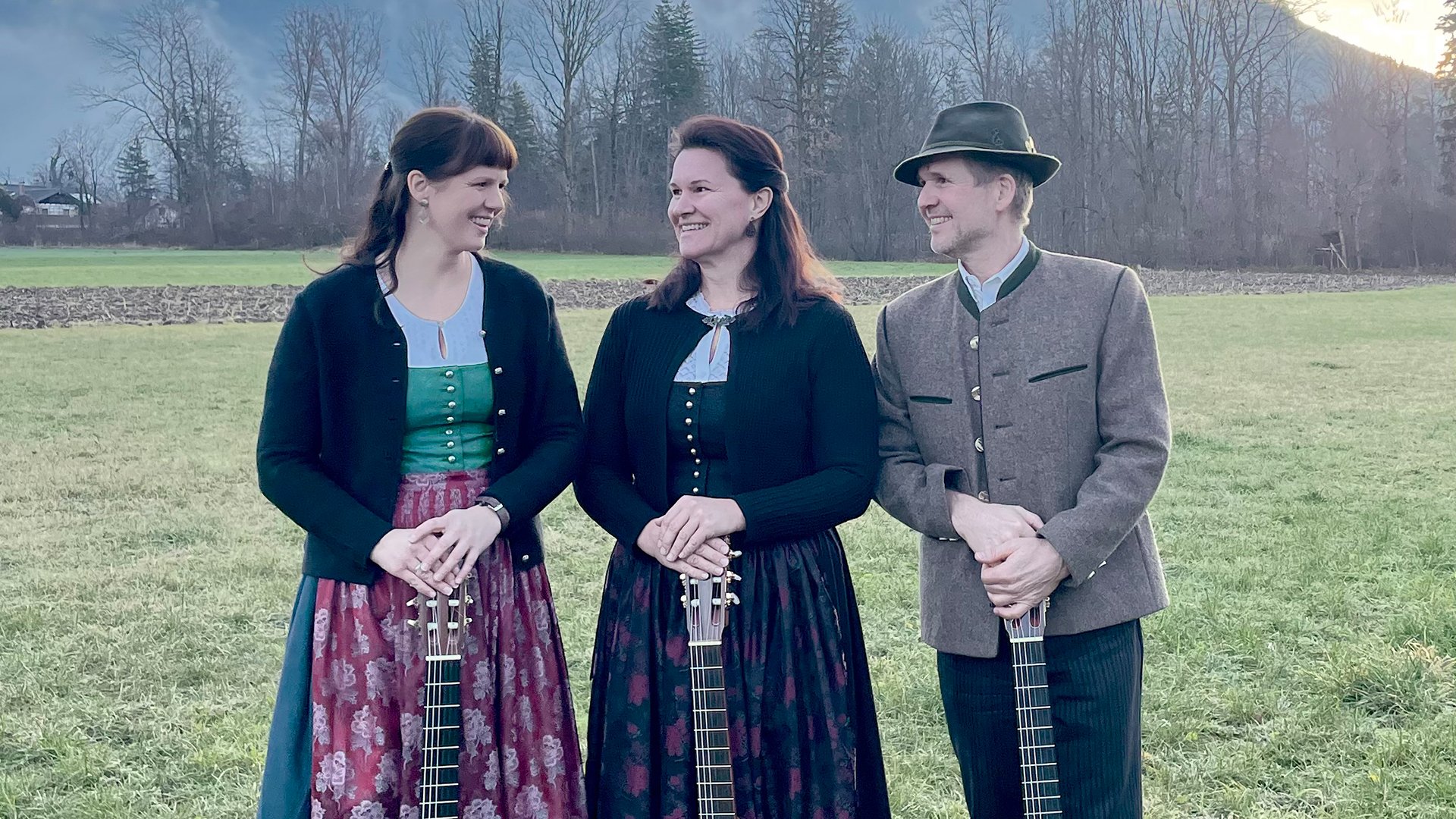 Drei Personen in Tracht mit je einer akustischen Gitarre auf einer Wiese. Zwei Frauen in Dirndln in Grün-, Rosa- und Schwarztönen, ein Mann mit graubrauner Trachtenjacke und Hut. Hintergrund mit Wiese, kahlen Bäumen und Bergen