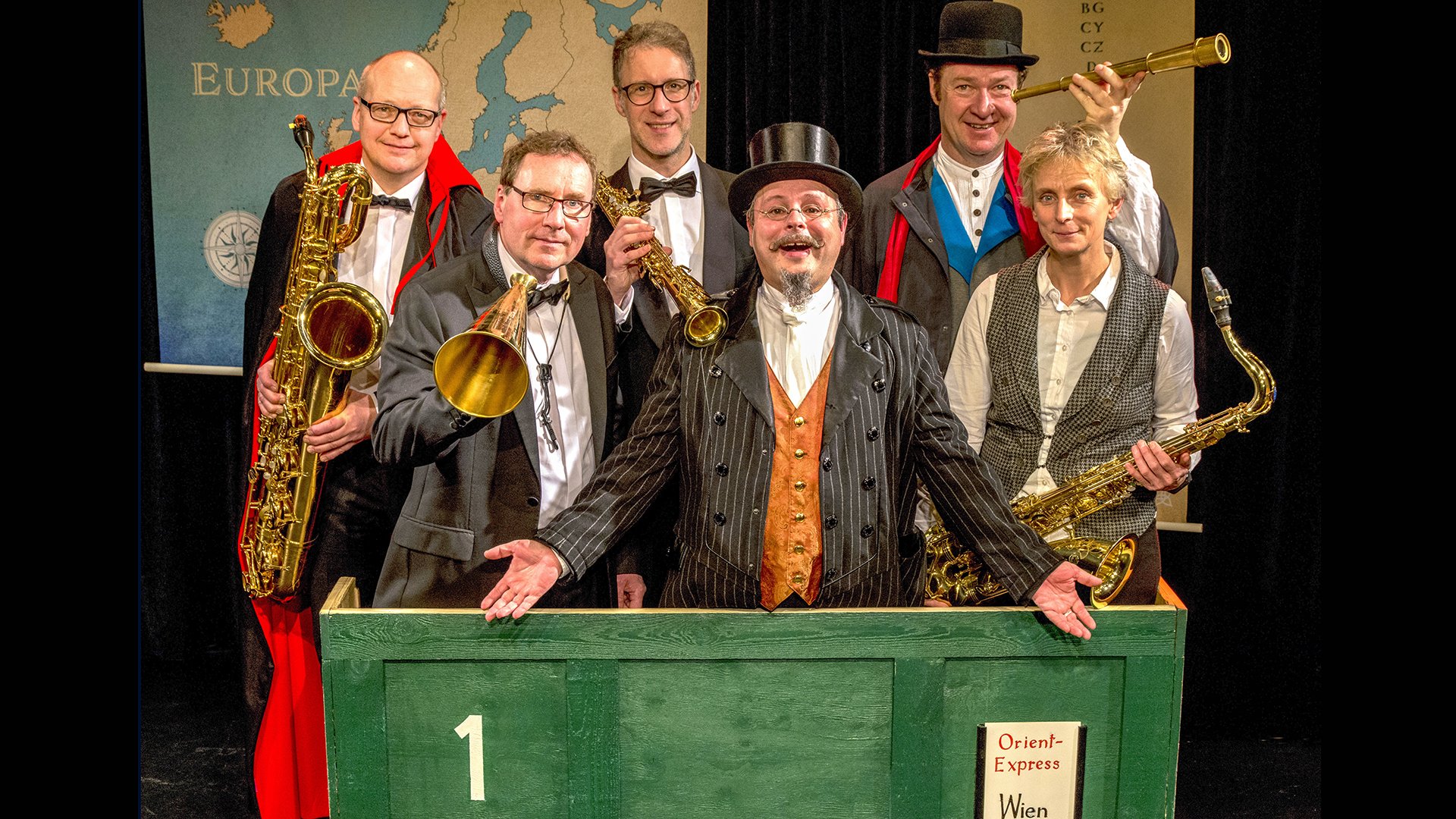 Sechs Personen in historischen und festlichen Kostümen posieren mit verschiedenen Blasinstrumenten und einem Fernrohr hinter einem grünen Zugabteil mit der Aufschrift Orient-Express Wien.
