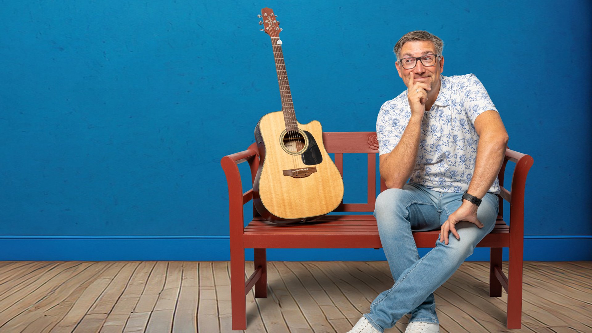 Foto für die Veranstaltung von Stefan OTTO – „So schee scho“ mit dem Künstler, der auf einer Bank sitzt, mit einer Gitarre vor blauem Hintergrund.