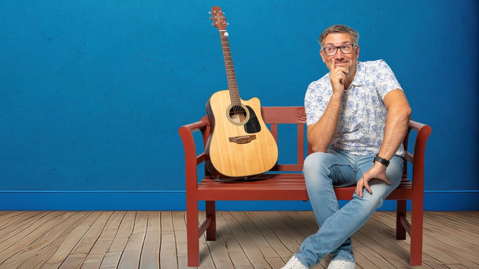 Foto für die Veranstaltung von Stefan OTTO – „So schee scho“ mit dem Künstler, der auf einer Bank sitzt, mit einer Gitarre vor blauem Hintergrund.