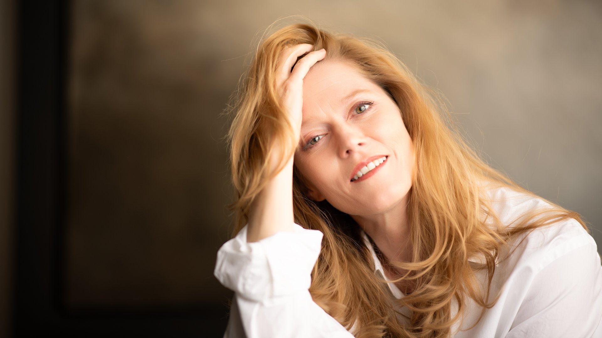Barbara Hannigan mit langen Haaren in heller Kleidung, den Kopf leicht geneigt und eine Hand im Haar, vor einem weich beleuchteten, neutralen Hintergrund