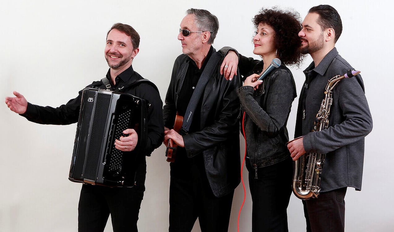 drei Künstler männlich stehen in einer Reihe mit einem Musikinstrument und eine Frau mit Mikrofon in der Hand vor weißem Hintergrund