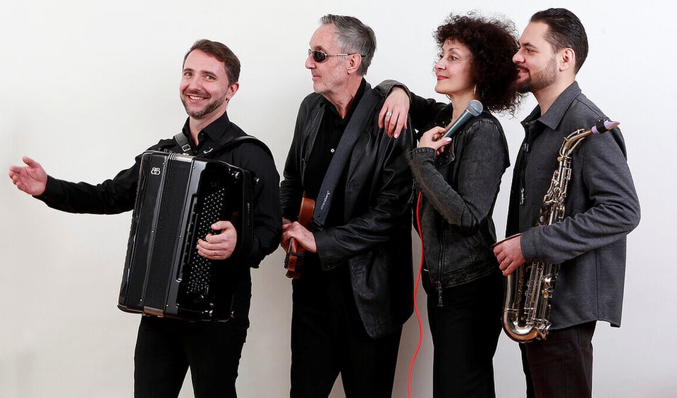 drei Künstler männlich stehen in einer Reihe mit einem Musikinstrument und eine Frau mit Mikrofon in der Hand vor weißem Hintergrund