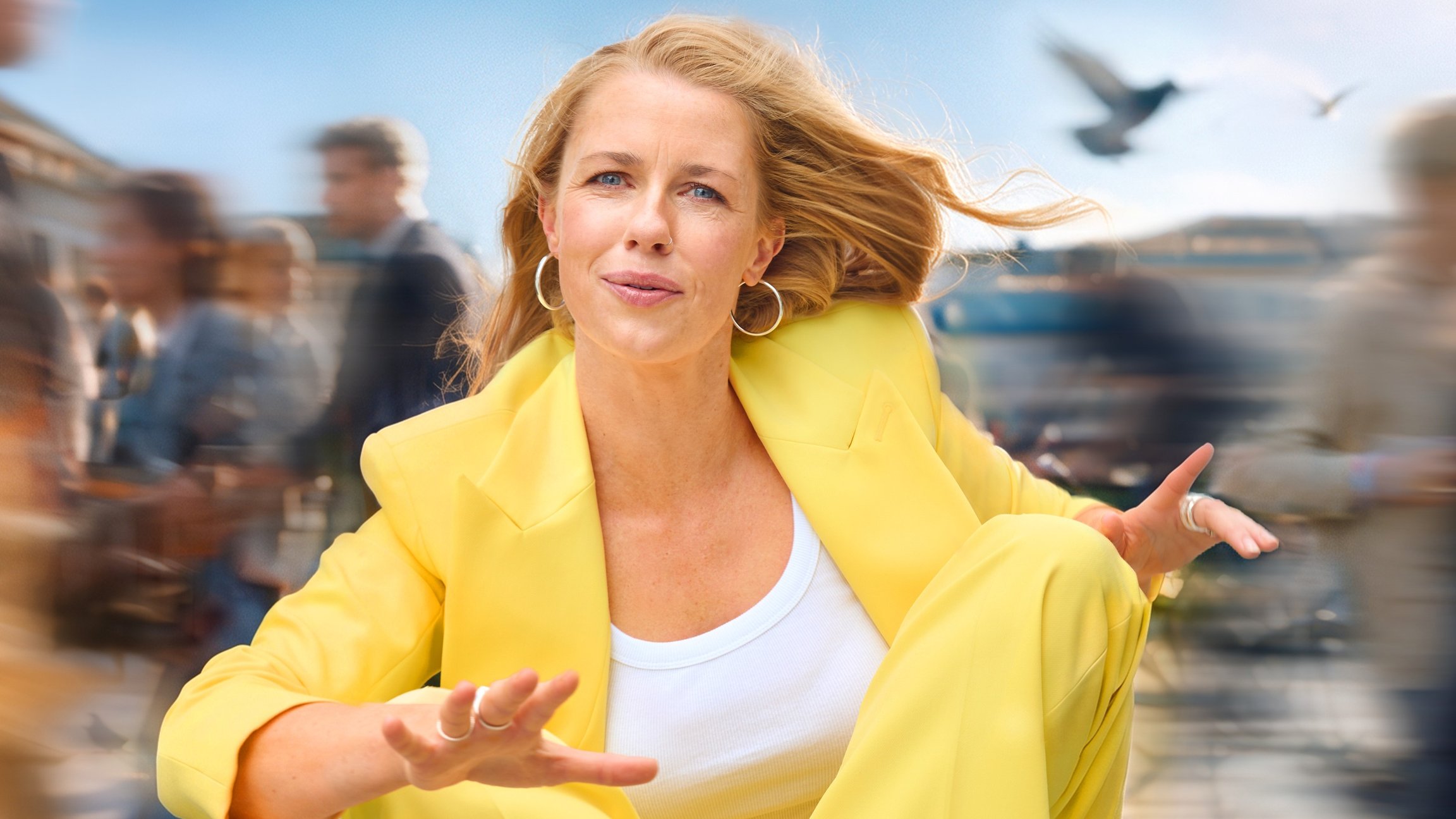 Christine Eixenberger in leuchtend gelbem Hosenanzug sitzt in dynamischer Pose im Freien. Im Hintergrund ziehen verschwommene Passantinnen und Passanten vorbei, während Wind durch die Haare weht und Vögel am Himmel zu sehen sind.