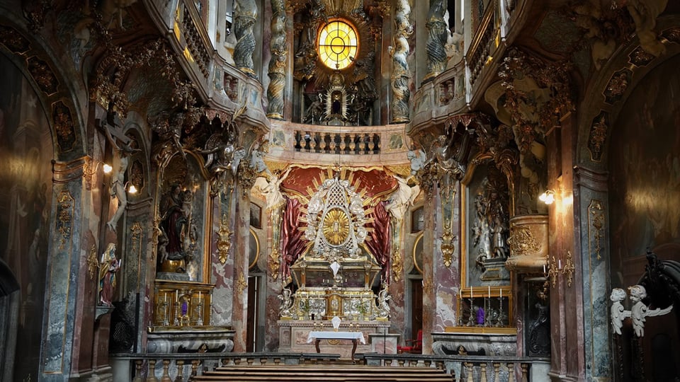 Innenraum der barocken Asamkirche mit reich verziertem Altar, farbigem Marmor, goldenen Ornamenten, Skulpturen und einem großen Rundfenster im oberen Bereich, frontal aufgenommen mit Blick auf den Hauptaltar.