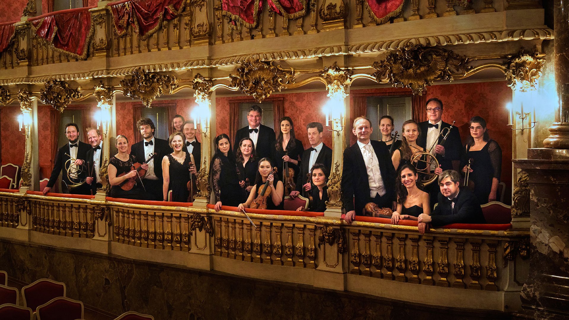 Gruppenfoto eines Orchesters auf einem reich verzierten Theaterbalkon mit goldenen Ornamenten und roten Vorhängen mehrere Personen in schwarzer Konzertkleidung halten Instrumente in einem historischen Saal