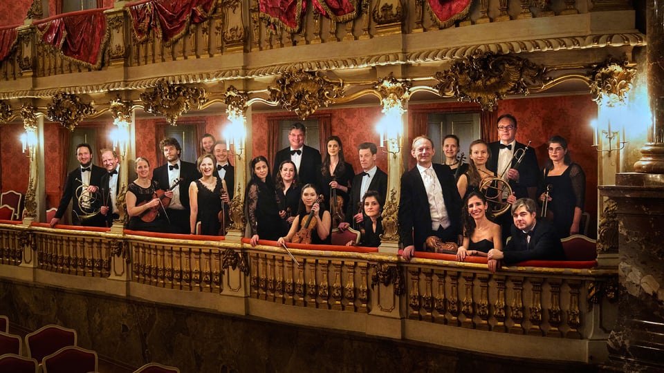 Gruppenfoto eines Orchesters auf einem reich verzierten Theaterbalkon mit goldenen Ornamenten und roten Vorhängen mehrere Personen in schwarzer Konzertkleidung halten Instrumente in einem historischen Saal