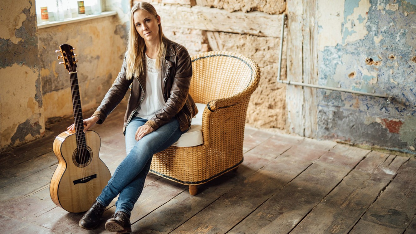 Die Künstlerin Julie Malia in Jeans und Lederjacke mit Akustikgitarre sitzt auf einem Korbstuhl in einem rustikalen Raum