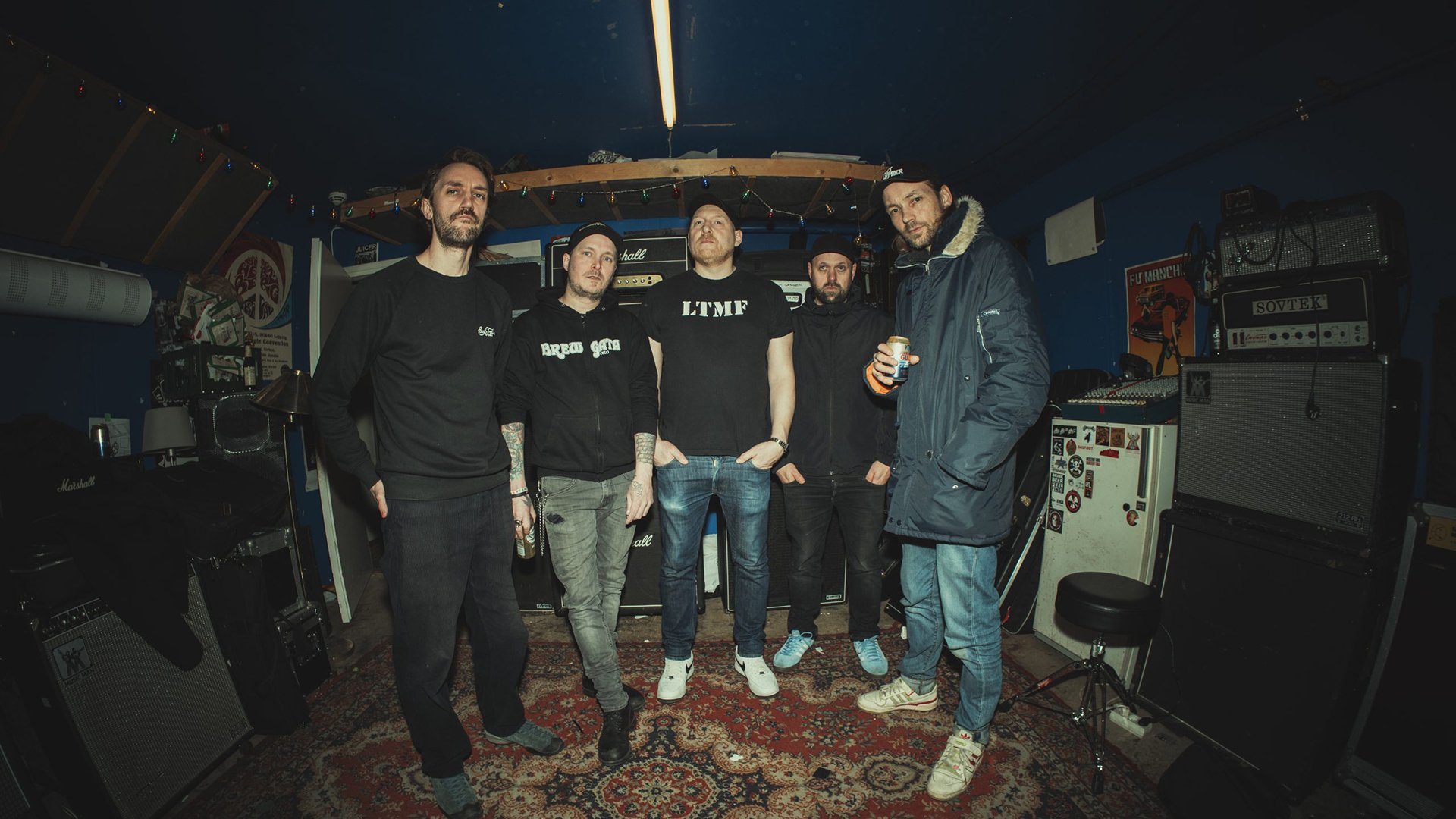 Bandfoto von The Good The Bad and The Zugly mit fünf Bandmitgliedern in dunkler Freizeitkleidung in einem Proberaum, umgeben von Verstärkern, Lautsprecherboxen und Musikequipment vor dunkelblauen Wänden.