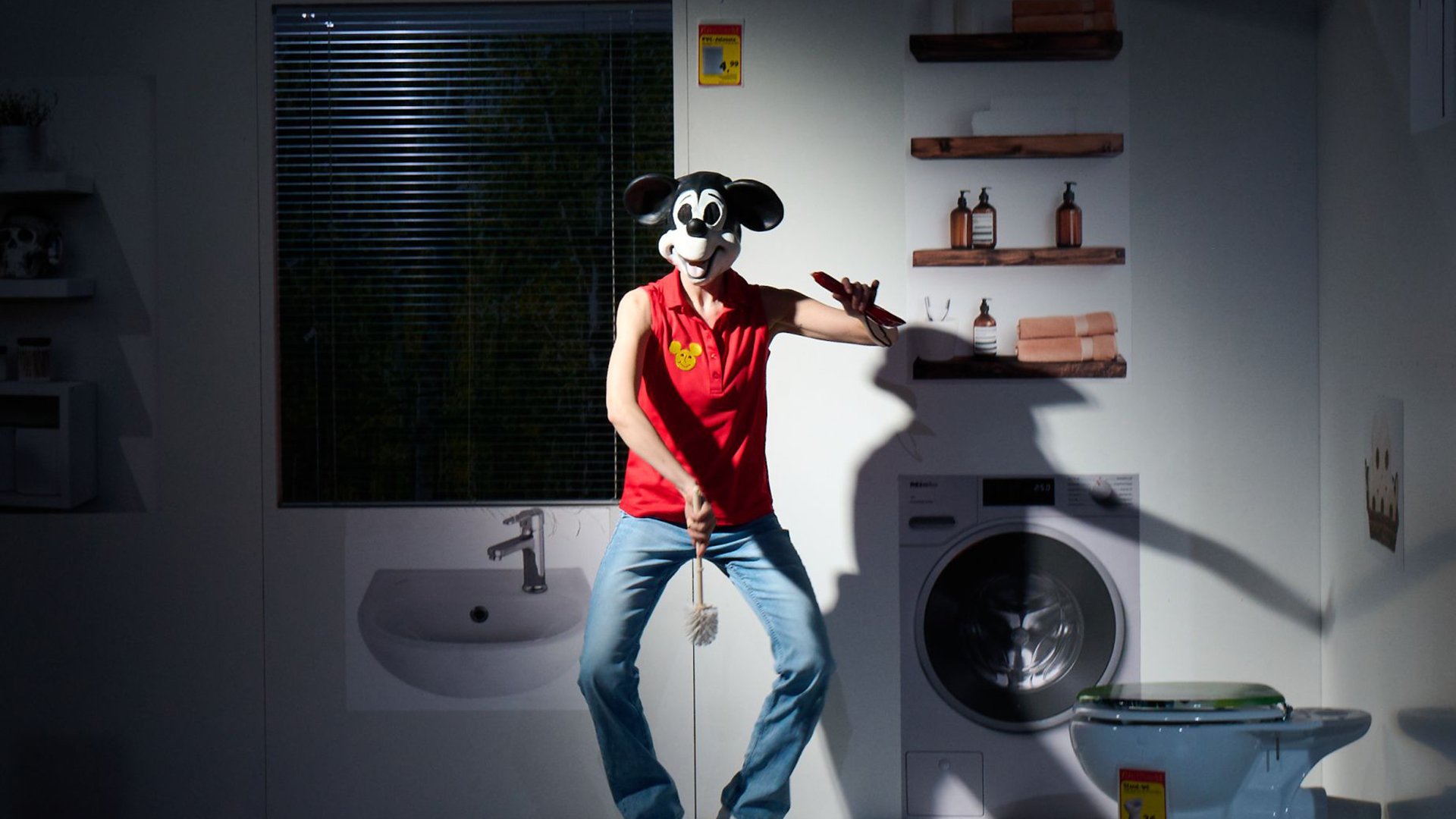 Person mit Mausmaske steht in einem Bühnenbild aus Badezimmer und Waschmaschine, mit ausgestrecktem Arm und Reinigungsutensil in der Hand
