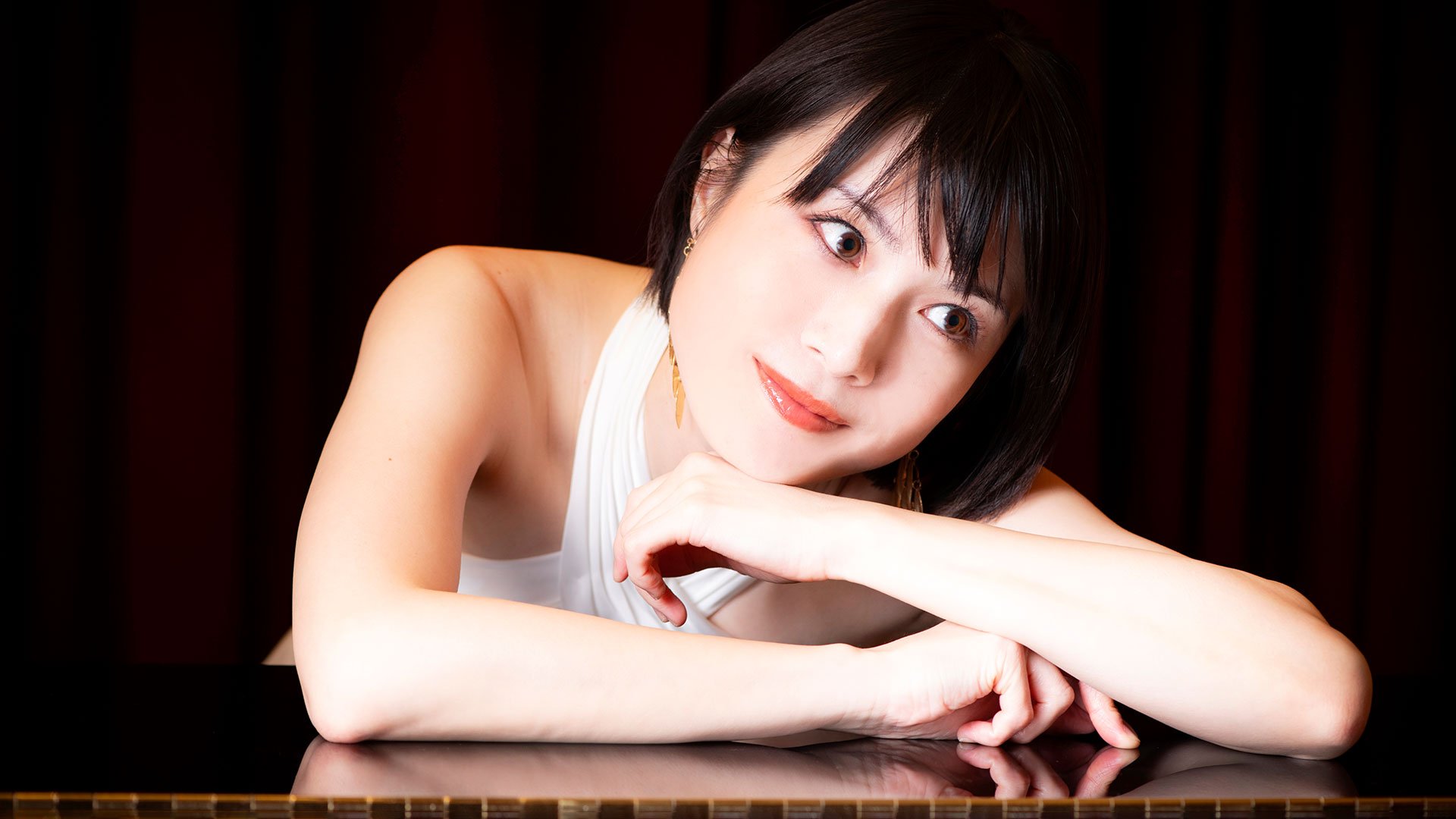 Foto für die Veranstaltung Shoko Kawasaki – Klavierkonzert mit der Künstlerin, die sich auf einen Tisch lehnt, vor schwarzem Hintergrund.