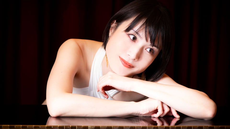 Foto für die Veranstaltung Shoko Kawasaki – Klavierkonzert mit der Künstlerin, die sich auf einen Tisch lehnt, vor schwarzem Hintergrund.