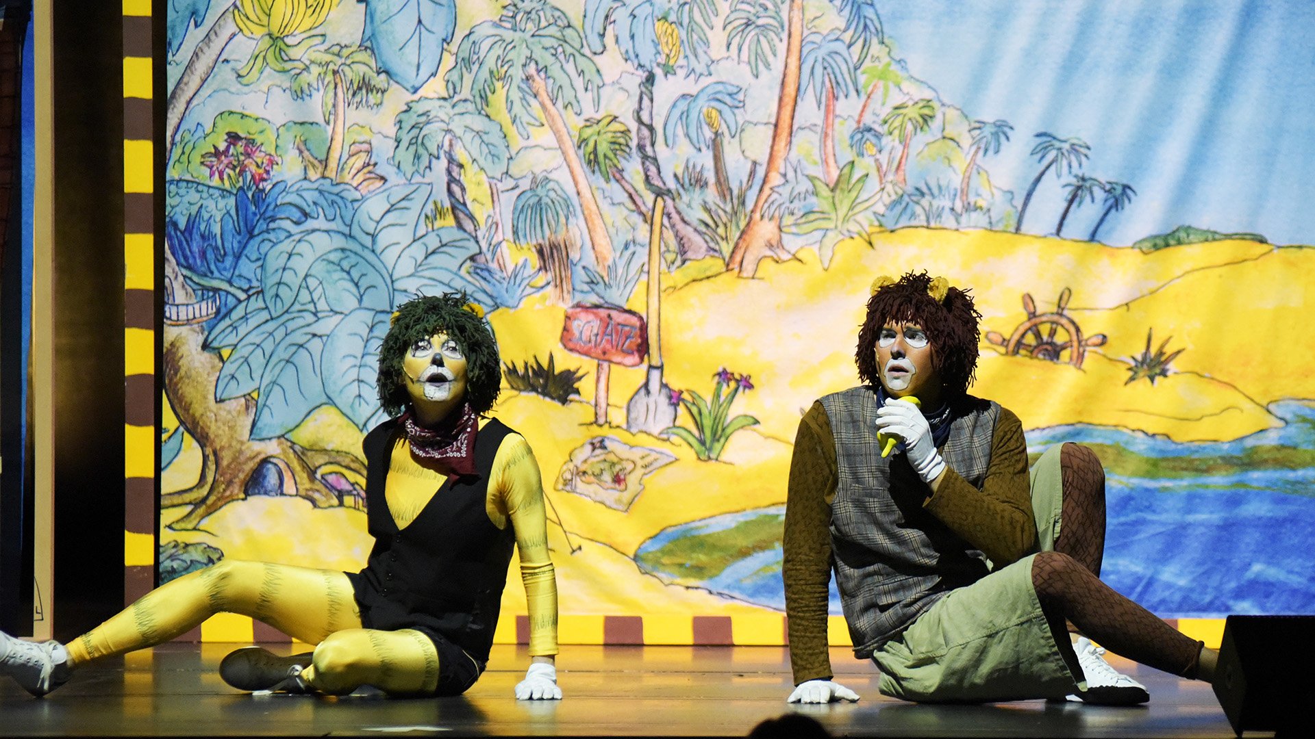Zwei als tierähnliche Figuren geschminkte Darsteller*innen sitzen auf einer Theaterbühne des Theaterstücks Janosch. Beide tragen Kostüme mit Felloptik und sitzen vor einem farbenfrohen, gemalten Bühnenbild, das eine tropische Landschaft mit Palmen, Pflanzen und einem Fluss zeigt. Eine der Figuren hält ein Mikrofon.