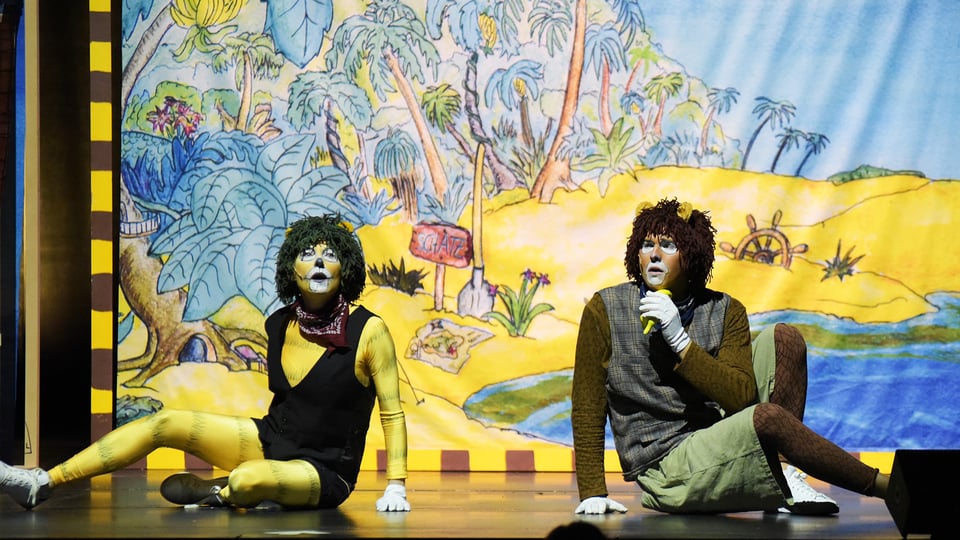 Zwei als tierähnliche Figuren geschminkte Darsteller*innen sitzen auf einer Theaterbühne des Theaterstücks Janosch. Beide tragen Kostüme mit Felloptik und sitzen vor einem farbenfrohen, gemalten Bühnenbild, das eine tropische Landschaft mit Palmen, Pflanzen und einem Fluss zeigt. Eine der Figuren hält ein Mikrofon.