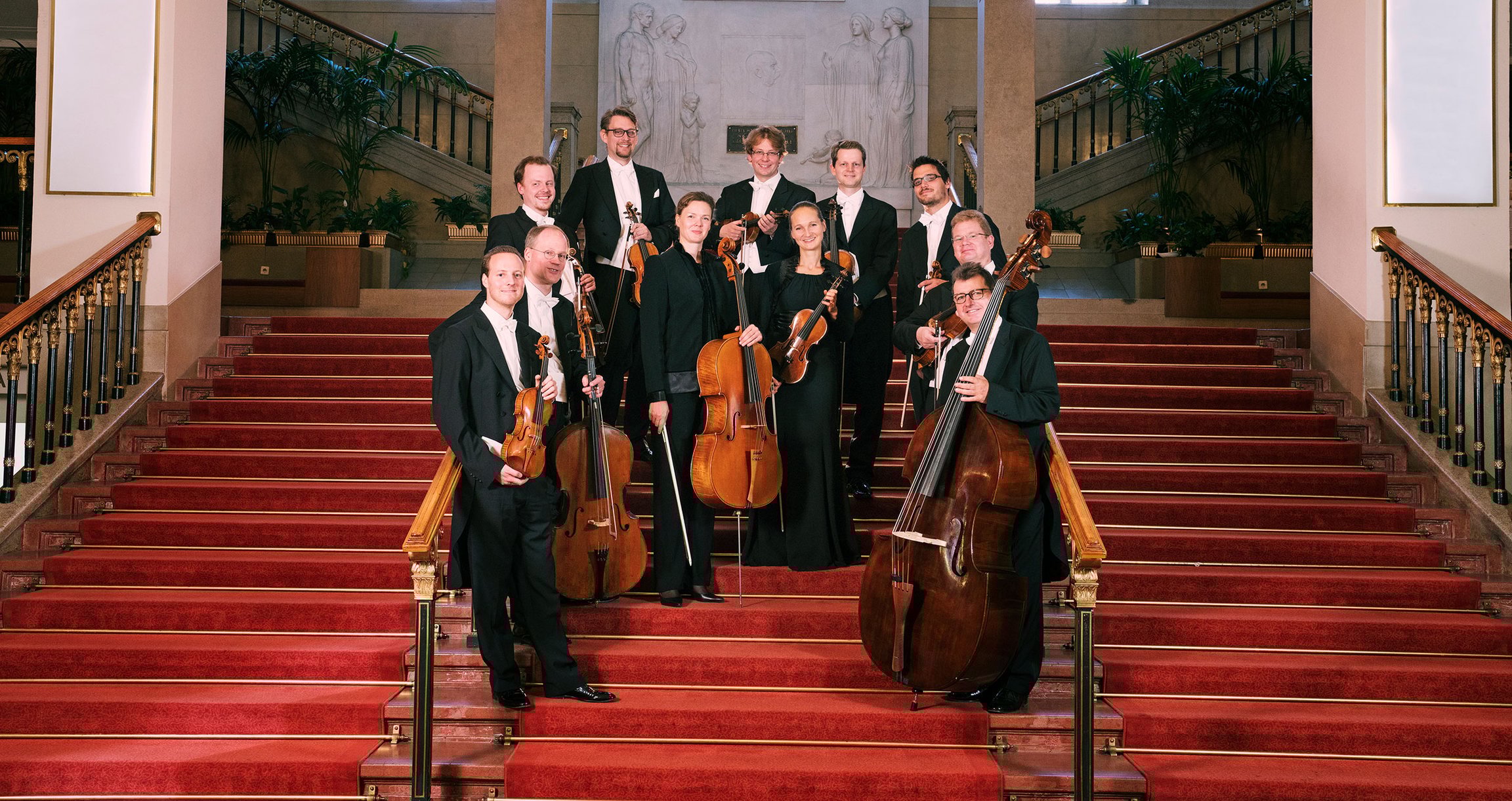 Orchester mit Streichinstrumenten in dunkler Kleidung auf roter Treppe