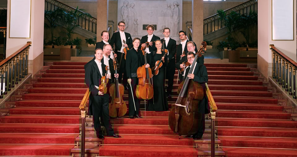 Orchester mit Streichinstrumenten in dunkler Kleidung auf roter Treppe