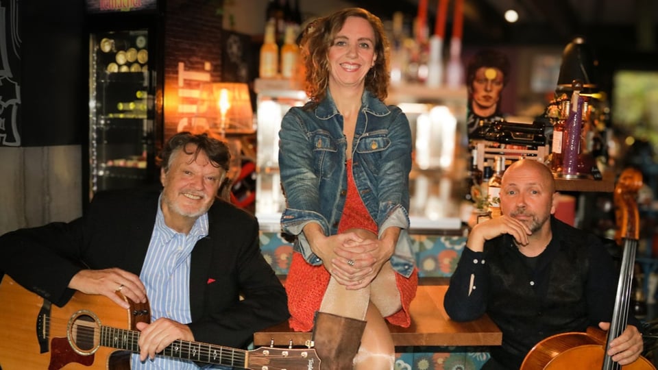Drei Personen in einem Café: eine sitzt auf dem Tresen, zwei sitzen davor mit Gitarre und Streichinstrument; im Hintergrund Bar und Musikinstrumente.