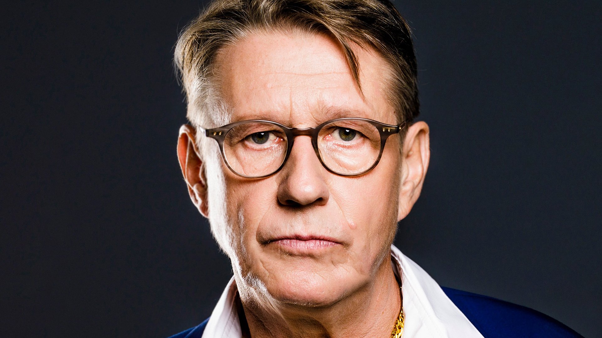 Ein Portrait von Andreas Rebers in weißem Hemd, goldener Kette und blauem Sakko vor dunklem Hintergrund