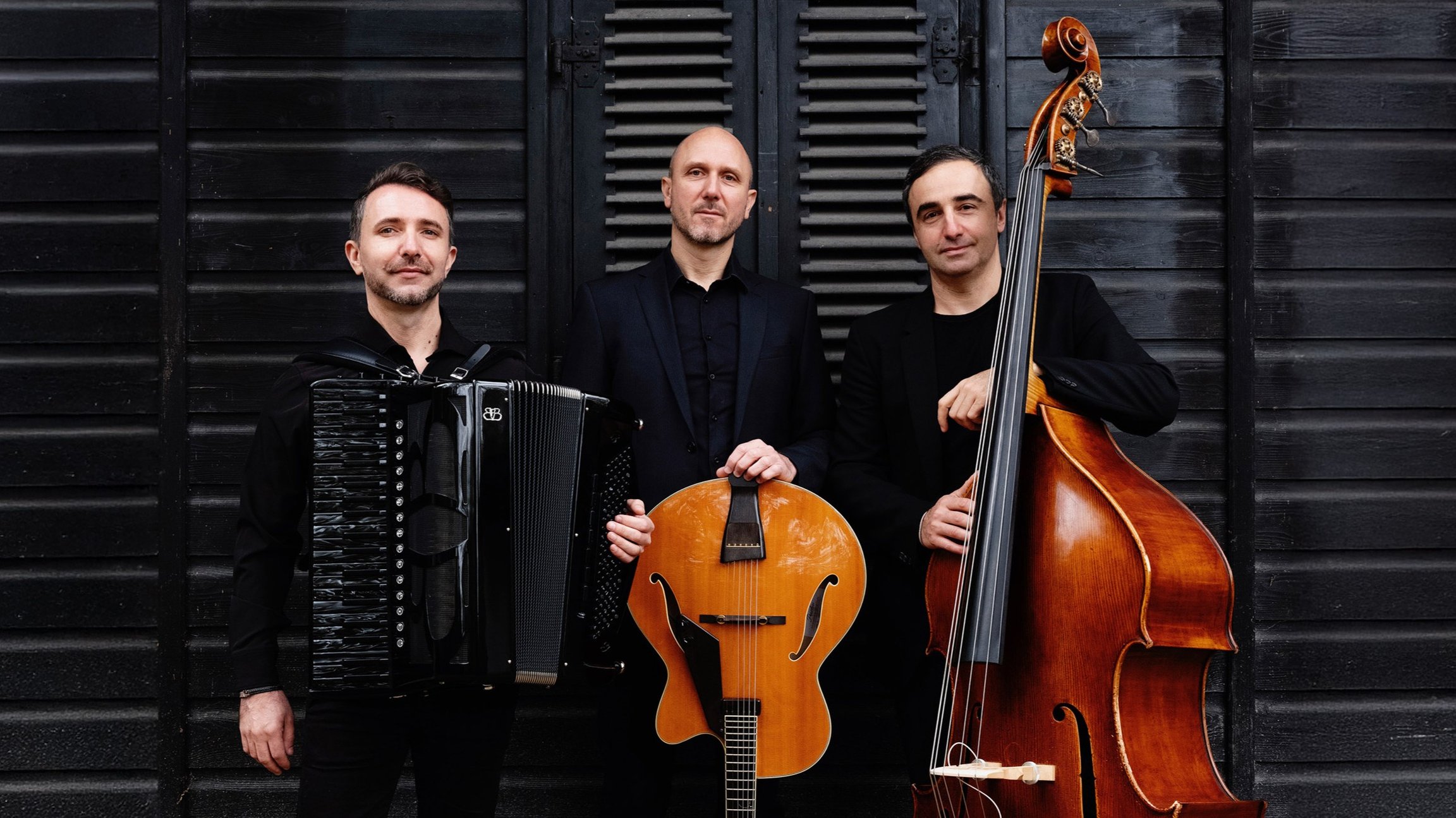 Die Band Acoustic Caravan vor einer schwarzen Holzwand. Drei Musiker in schwarzer Kleidung mit Akkordeon, Gitarre und Kontrabass. Die Instrumente setzen braune und schwarze Farbakzente.
