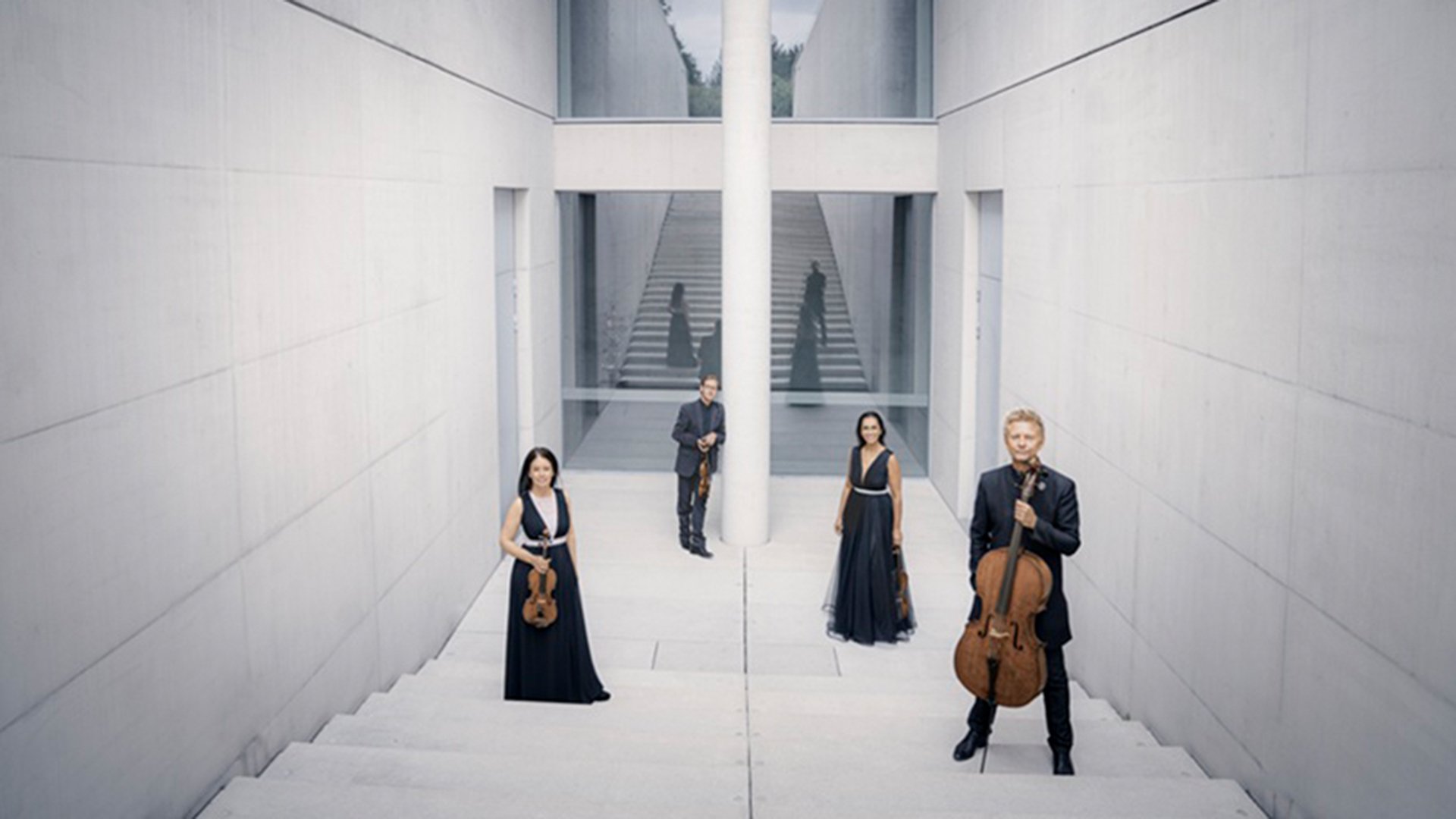 Streichquartett in einem modernen, hellen Treppenhaus. Die vier Ensemblemitglieder stehen mit ihren Streichinstrumenten auf einer breiten Treppenanlage vor klarer Architektur mit Treppen, Glasflächen und zentraler Säule