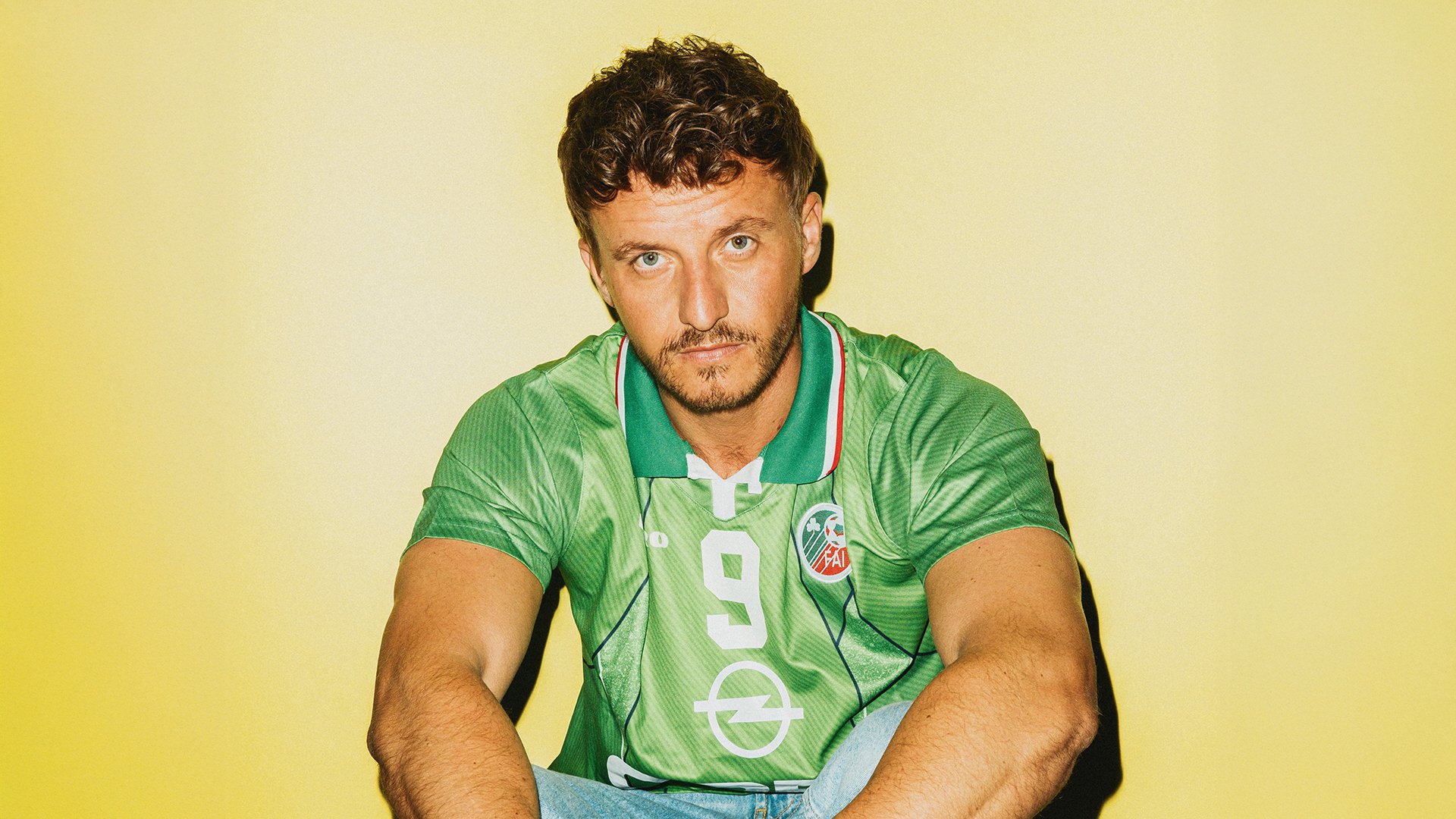 Porträtfoto von Copa TS in grünem Fußballtrikot mit der Nummer 9 vor gelbem Hintergrund, auf dem Boden sitzend und direkt zur Kamera blickend, mit Schattenwurf an der Wand hinter ihm.