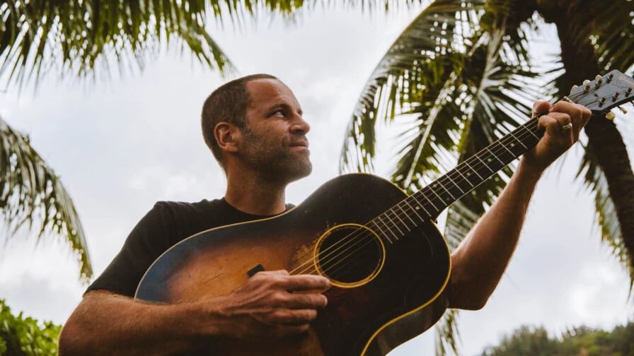 Jack Johnson mit Gitarre im Freien unter Palmen zur Seite blickend 