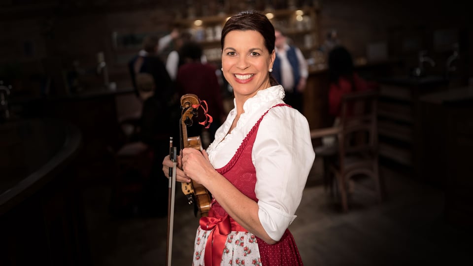 Person in traditioneller Tracht mit rotem Dirndl steht in einem Wirtshaus und hält eine Violine, im Hintergrund unscharf Gäste und Holzmobiliar in gemütlicher Atmosphäre