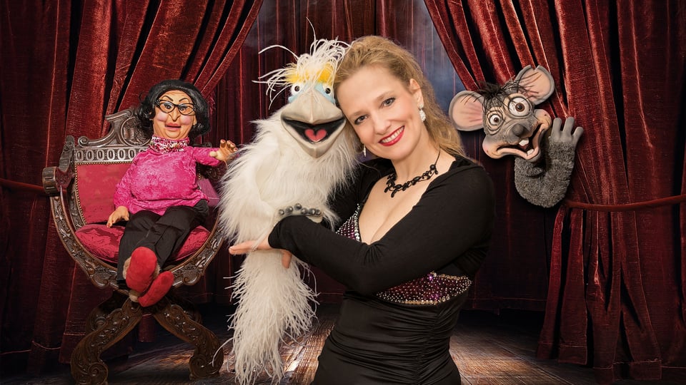 Bühnenbild mit rotem Vorhang, Murzarella in schwarzem Kleid präsentiert drei Handpuppen (Vogel, Maus, ältere Figur) und eine auf einem Sessel sitzende Bauchrednerpuppe