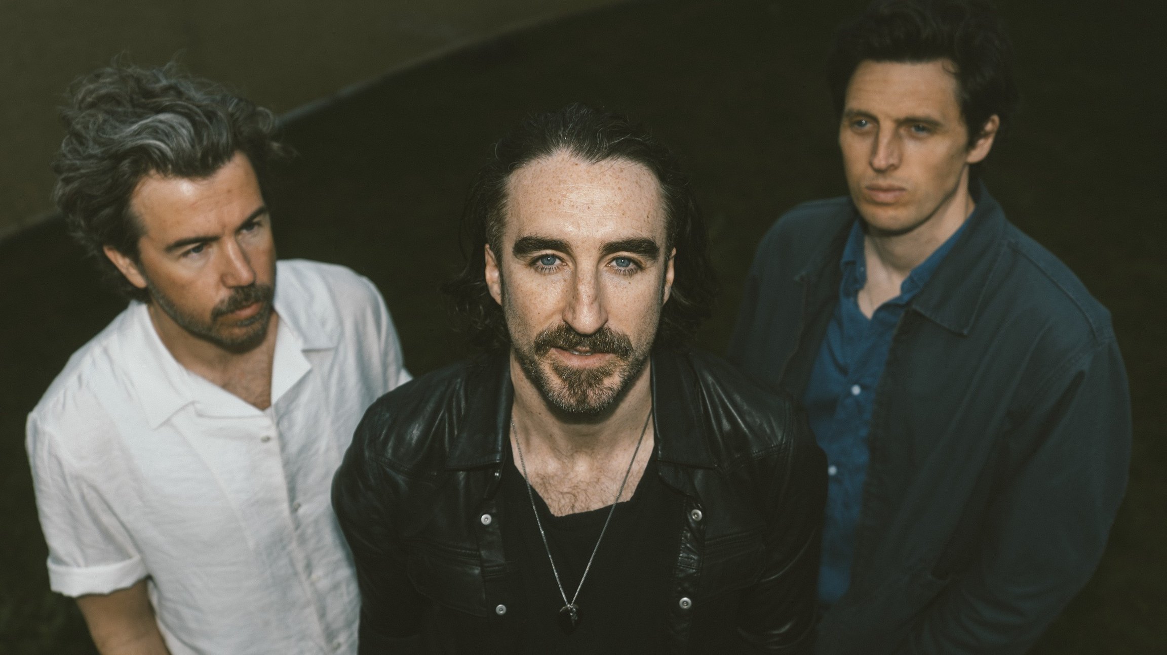 Die dreiköpfige Band The Coronas stehen zusammen, die mittlere Person blickt in die Kamera, vor dunklem Hintergrund