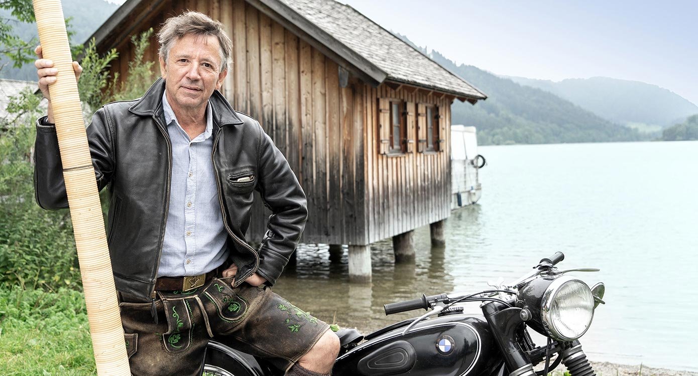 Ein Foto von einem Mann, der auf einem Motorrad sitzt, im Hintergrund eine Hütte und ein See.