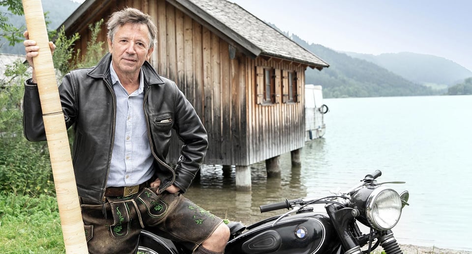 Ein Foto von einem Mann, der auf einem Motorrad sitzt, im Hintergrund eine Hütte und ein See.