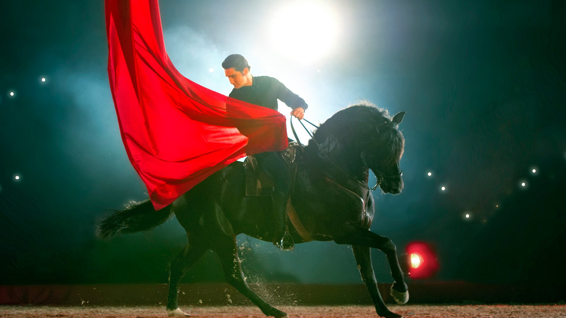 Reiterische Szene auf einer beleuchteten Bühne: Eine Person reitet ein schwarzes Pferd, während ein großes rotes Tuch im Licht weht, vor dunklem Hintergrund mit punktförmigen Lichtern