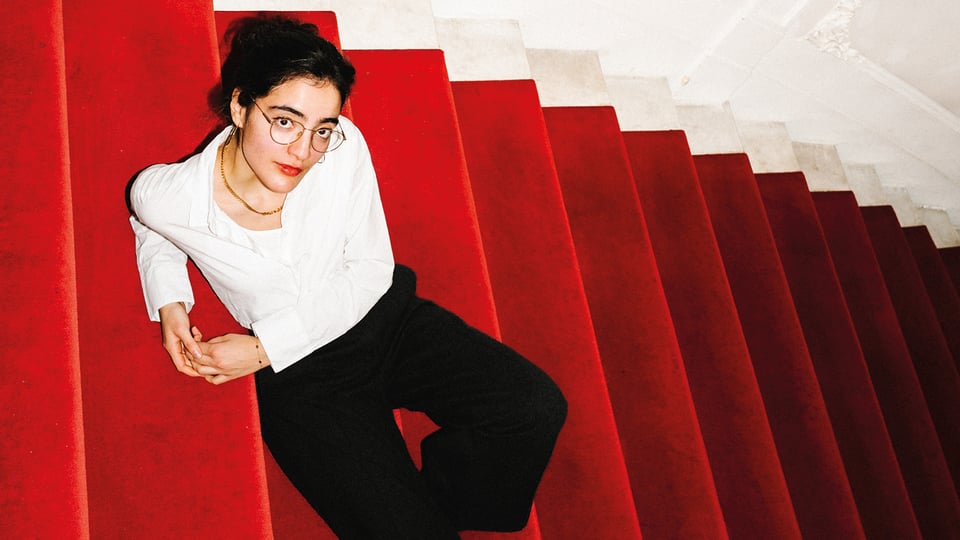 Eine Person sitzt auf einer roten, mit Teppich belegten Treppe. Sie trägt eine weiße Bluse, eine schwarze Hose und eine Brille. Die Treppe verläuft diagonal durch das Bild, der obere Bereich ist in hellen Weiß- und Cremetönen gestaltet.