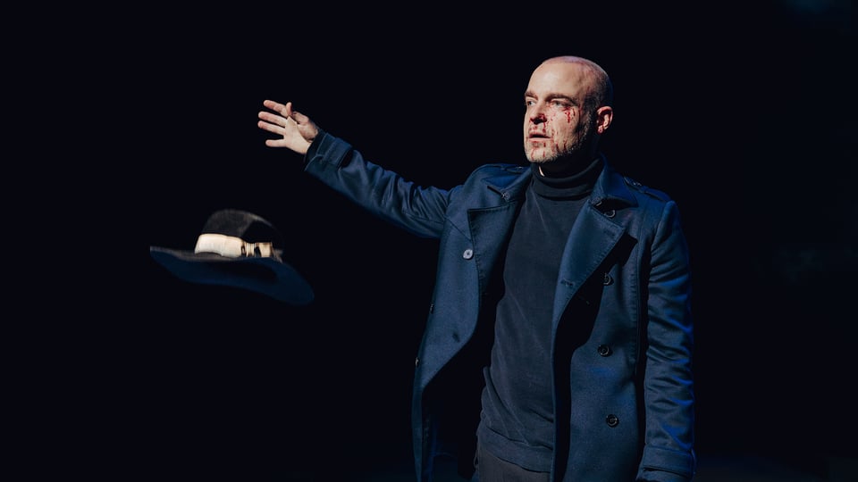 Der Darsteller in dunkelblauem Anzug und mit blutverschmiertem Gesicht streckt seinen Arm aus